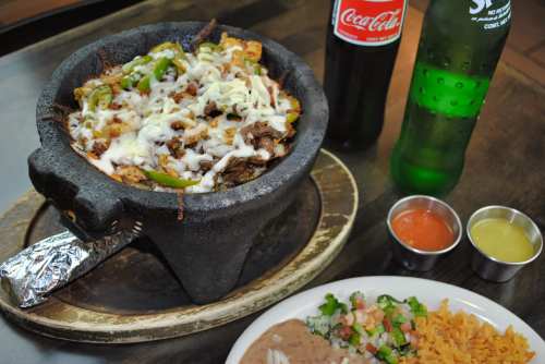 A sizzling stone bowl filled with meat, peppers, and cheese, accompanied by drinks and sides of rice and salsa.