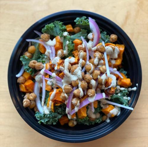 A vibrant salad with kale, roasted sweet potatoes, chickpeas, red onion, and a creamy dressing in a black bowl.