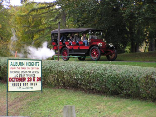 A vintage car with passengers drives past a sign for Auburn Heights, promoting a steam car museum event.