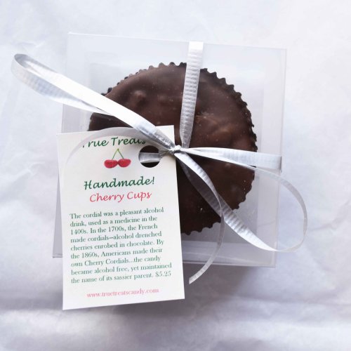 A chocolate-covered cherry cup in a clear box, tied with a ribbon and a tag explaining its history.