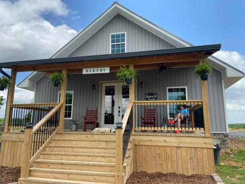 A charming gray bakery with a wooden porch, featuring rocking chairs and potted plants under a blue sky.