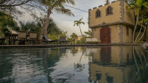 A serene pool scene featuring a unique stone building, palm trees, and lounge chairs under a cloudy sky.