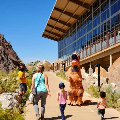 A person in a dinosaur costume walks along a path with children near a modern building and rocky landscape.
