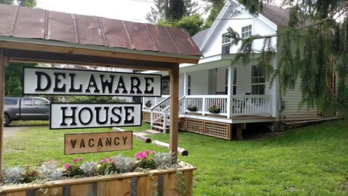 A sign reading "Delaware House" with "Vacancy" below, in front of a charming white house with a porch and greenery.