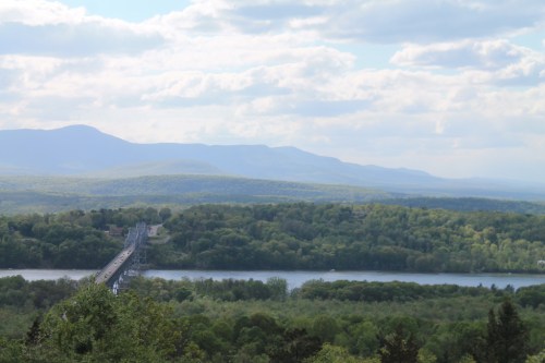 A scenic view of a bridge spanning a river, surrounded by lush green trees and distant mountains under a cloudy sky.