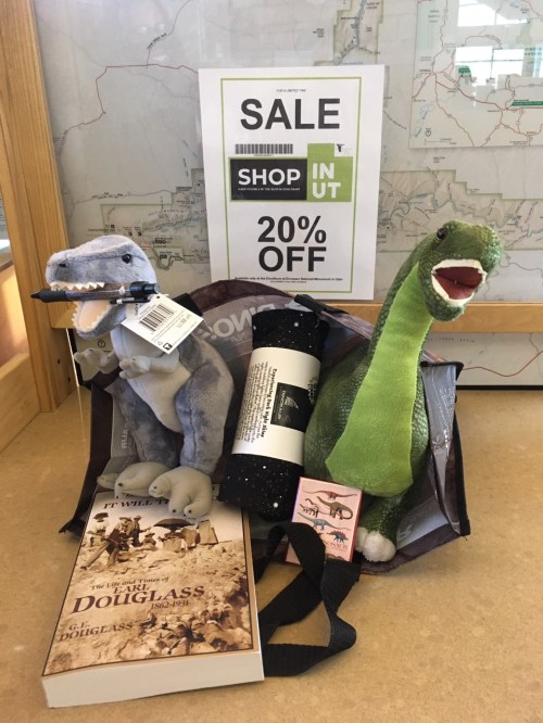 Stuffed dinosaurs, a book, and a rolled-up item in a bag, with a sign advertising a 20% off sale.