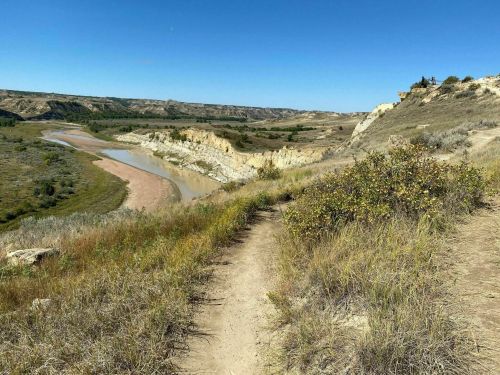 A winding dirt path leads through grassy hills overlooking a river and rugged terrain under a clear blue sky.