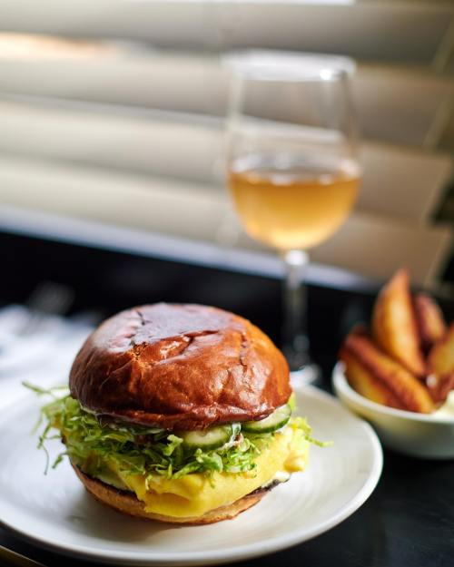 A close-up of a burger with lettuce and pickles on a plate, accompanied by a glass of wine and side of fries.