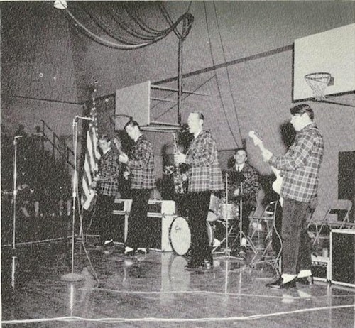 A band performs on stage in a gymnasium, wearing plaid shirts, with instruments and a microphone set up.