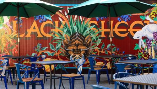 Colorful mural featuring a stylized face and tropical plants, with outdoor tables and umbrellas in a vibrant setting.