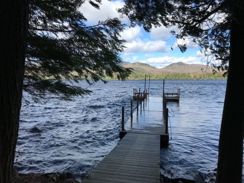 A wooden dock extends into a lake, surrounded by trees and mountains under a partly cloudy sky.