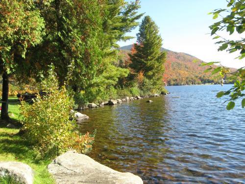 A serene lakeside scene with colorful autumn trees reflecting in the calm water under a clear blue sky.