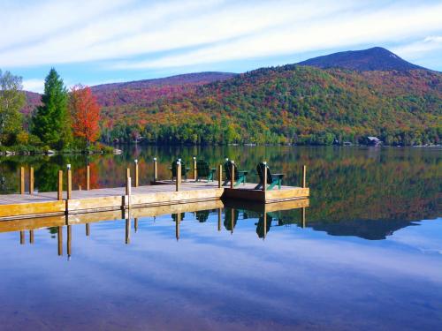 A serene lake scene with a wooden dock, chairs, and colorful autumn foliage reflecting on the calm water.