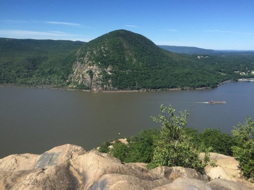 A scenic view of a green mountain rising above a river, with a boat navigating the water under a clear blue sky.