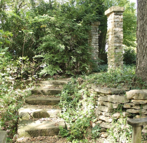 Stone steps lead up through lush greenery to ancient stone pillars in a serene garden setting.