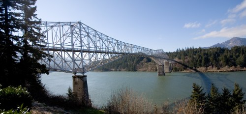 A large metal bridge spans a river, surrounded by trees and mountains under a clear blue sky.