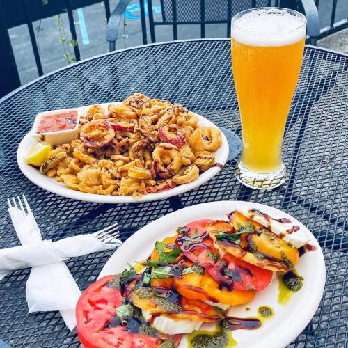 A plate of fried calamari with dipping sauce and a colorful tomato salad, alongside a glass of beer on a table.