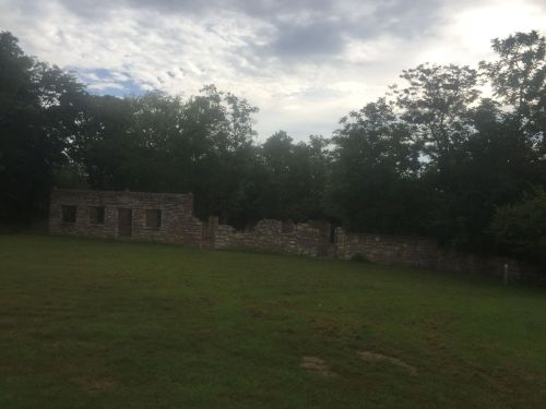 Ruins of a stone building surrounded by trees and grass under a cloudy sky.