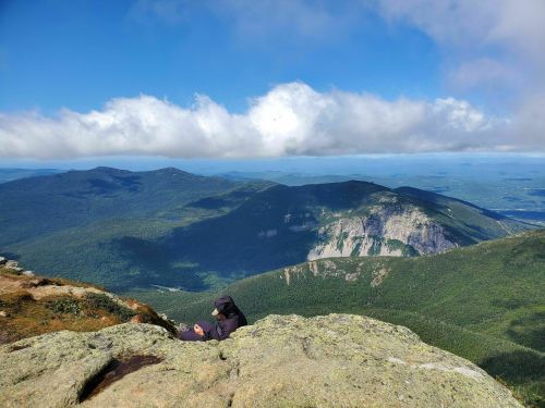 A person sits on a rocky ledge, overlooking a vast green mountain landscape under a partly cloudy sky.