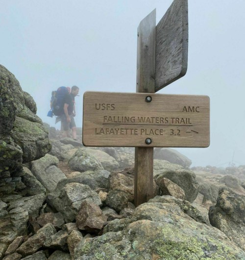 A wooden trail sign points to Falling Waters Trail, with hikers and rocky terrain in a foggy setting.