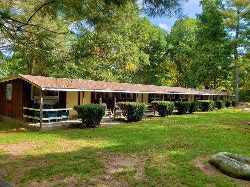 A long, wooden cabin with a porch, surrounded by green grass and trees under a clear blue sky.
