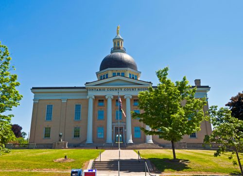 Front view of the Ontario County Courthouse, featuring a dome and flag, surrounded by trees under a clear blue sky.