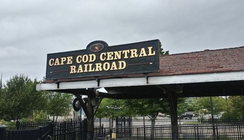Sign for Cape Cod Central Railroad under a cloudy sky, with trees and a fence in the background.