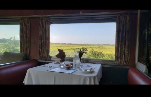 A dining table set with a white tablecloth and glassware, with a scenic view of green fields through the window.