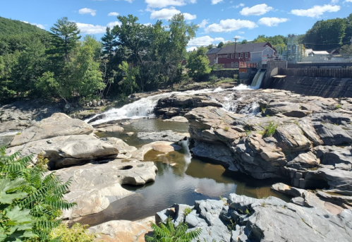 A scenic view of a river with rocks, a waterfall, and a building in the background under a blue sky with clouds.