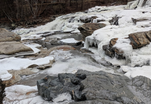 Frozen waterfall and rocky landscape with ice and snow, surrounded by bare trees in a winter setting.