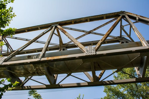 A weathered metal bridge structure with a large cylindrical pipe underneath, set against a clear blue sky.