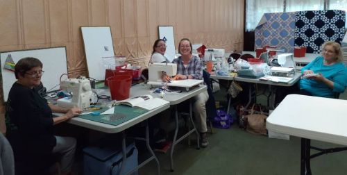 A group of women happily sewing at tables in a craft room, surrounded by sewing machines and materials.