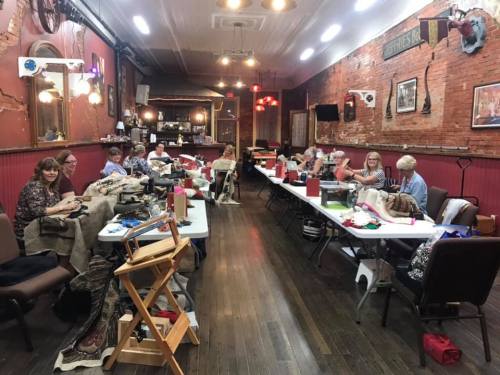 A group of people sewing at tables in a rustic room with brick walls and warm lighting.