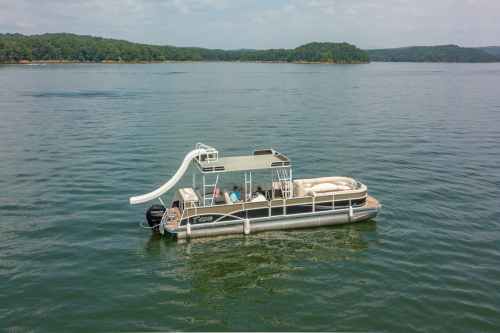 A pontoon boat with a slide is anchored on calm water, surrounded by green hills under a partly cloudy sky.