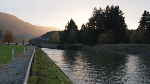 A serene river scene at sunset, surrounded by trees and mountains, with a pathway and bridge in the background.