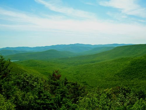 A panoramic view of lush green mountains under a clear blue sky, showcasing rolling hills and distant peaks.