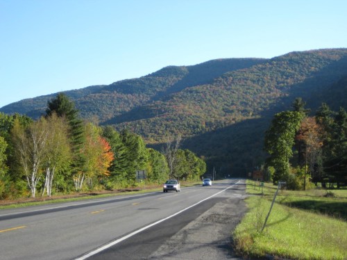 A scenic highway lined with trees, showcasing vibrant fall foliage against a backdrop of mountains under a clear blue sky.
