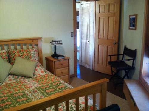 Cozy bedroom with floral bedding, wooden furniture, a bedside lamp, and a chair near an open door leading to another room.