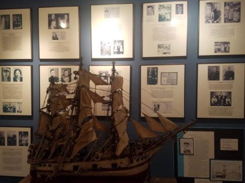 A model ship displayed in front of a wall featuring framed photographs and informational plaques.