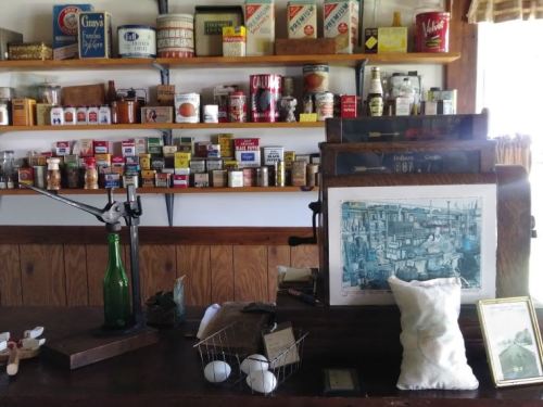 A vintage display featuring jars, cans, and a wooden counter with a scale, eggs, and an artwork in a rustic setting.