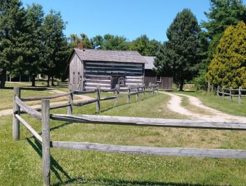 A rustic log cabin surrounded by a wooden fence, with a dirt path leading to it, set against a clear blue sky.