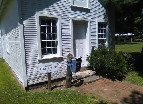 Historic white building with a sign reading "Lyme Post Office 1824" and a blue mailbox in front. Green grass surrounds it.