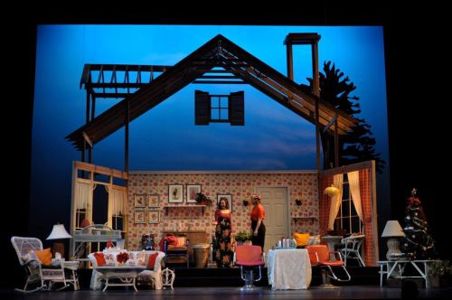 A stage set depicting a colorful, partially constructed house interior with furniture and decorations, under a blue sky backdrop.