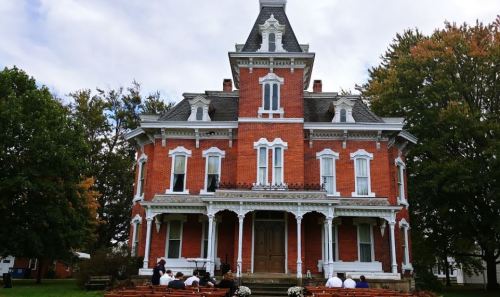 A large, red brick Victorian house with a steep roof and ornate details, surrounded by trees and people seated outside.