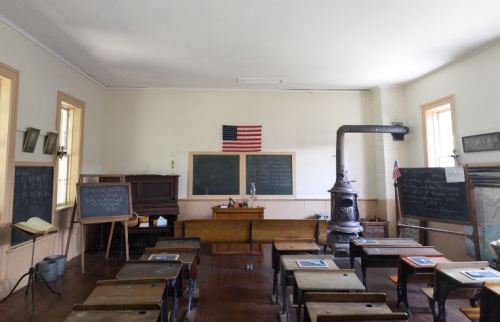 A vintage classroom with wooden desks, chalkboards, an American flag, and a potbelly stove.