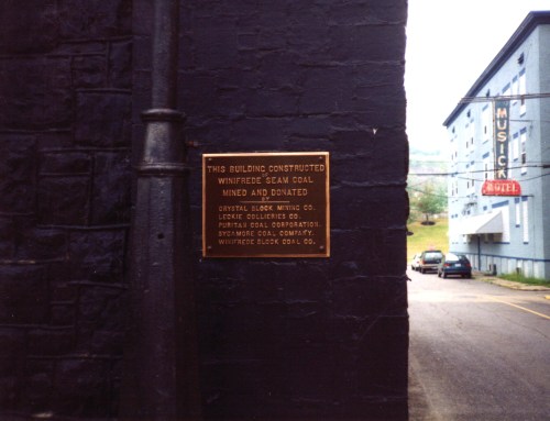 Plaque on a black wall, noting the building's construction from Winifrede seam coal, with a hotel visible in the background.