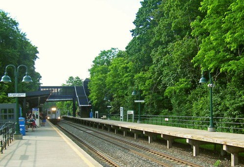 A train station platform surrounded by lush green trees, with a train approaching in the distance.