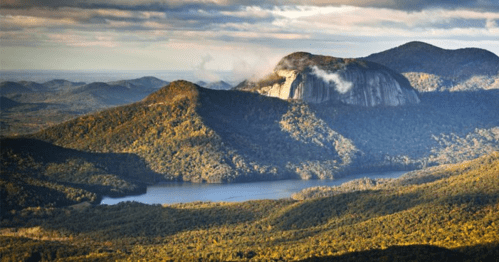 A panoramic view of a mountainous landscape with a prominent rock formation and a serene lake below, surrounded by autumn foliage.