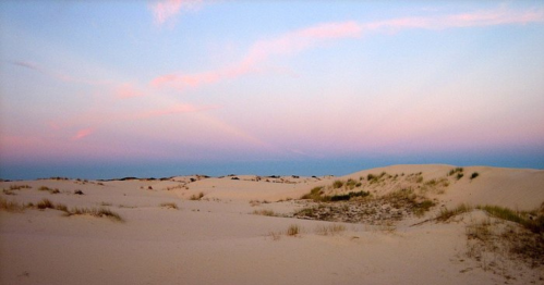 A serene landscape of sand dunes under a pastel sky at dusk, with gentle grasses peeking through the sand.