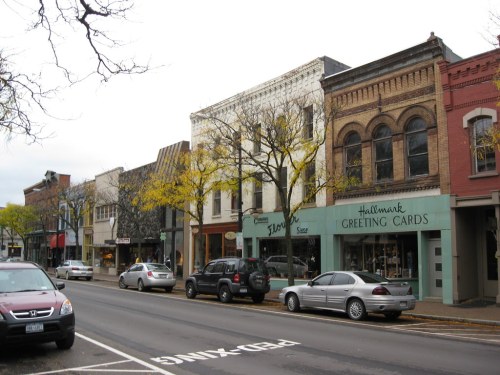 A quaint street lined with shops, including a Hallmark store, and trees with yellow leaves in a small town.